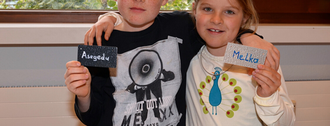 Ein kleiner Junge und ein kleines Mädchen halten zwei Namensschilder in die Höhe. Sie halten sich gegenseitig um die Schulter und grinsen.
