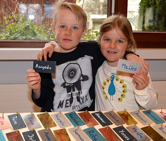 Ein kleiner Junge und ein kleines Mädchen halten zwei Namensschilder in die Höhe. Sie halten sich gegenseitig um die Schulter und grinsen.