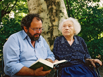 Ein Mann mittleren Alters mit einer Brille, Vollbart, schwarzen Haaren und einem blauen Hemd ließt aus einem Buch einer Dame im hohen Alter vor. Die Dame hat weiße Haare, ist dunkel gekleidet und sitzt in einem Rollstuhl. 