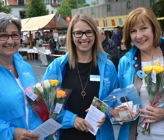 Drei Frauen mittleren Alters tragen alle dieselbe Regenjacke. Sie halten eine Spendenkasse und Rosen zum verkaufen in der Hand. 
