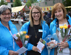 Drei Frauen mittleren Alters tragen alle dieselbe Regenjacke. Sie halten eine Spendenkasse und Rosen zum verkaufen in der Hand. 