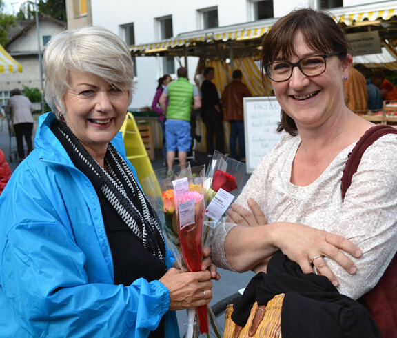 Zwei Frauen mittleren Alters lächeln in die Kamera. Eine Frau hält verschiedene Rosen in der Hand.
