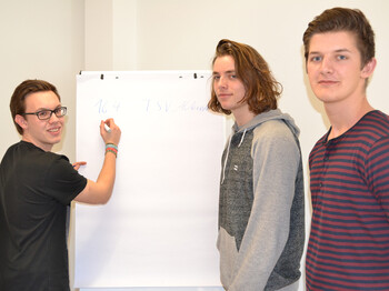 Drei junge Erwachsene stehen vor einem Flipchart. Einer davon schreibt etwas auf eine Seite des Flipchartes. Die anderen beiden schauen in die Kamera.