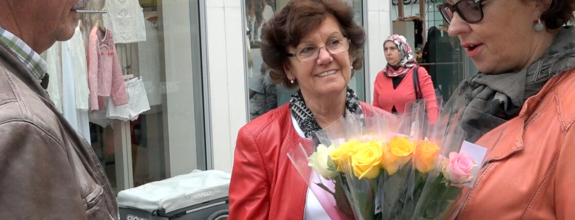 Drei Menschen treffen sich in der Stadt in Dornbirn. Eine Frau hält mehrere bunte Rosen in der Hand. Im Hintergrund sieht man ein Einkaufsgeschäft.