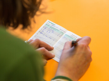 Eine Person mit grünem Oberteil notiert etwas in einem kleinen Heftchen mit der Aufschrift "Trink-Tagebuch".
