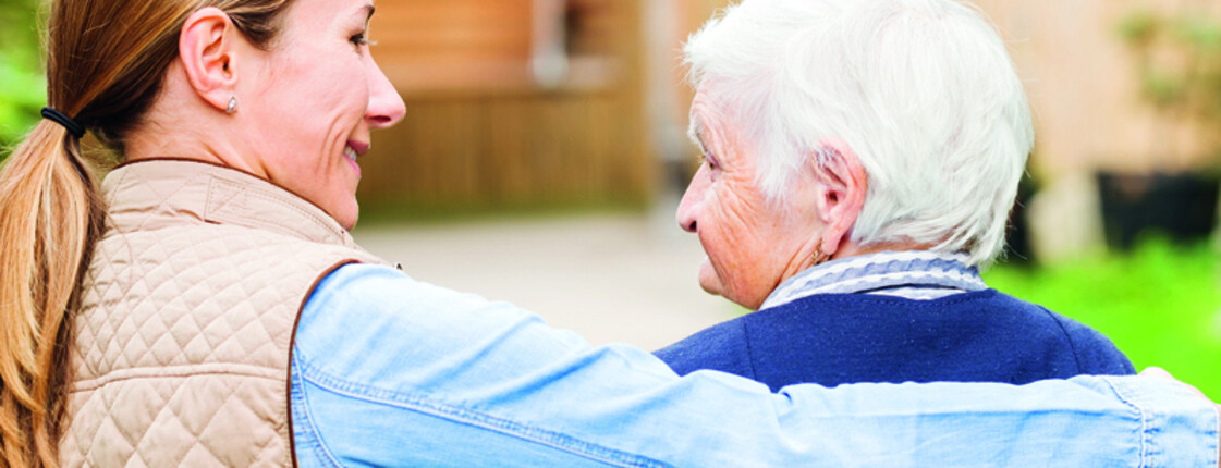Eine Frau mittleren Alters mit braunen langen Haaren umarmt eine ältere Dame, die kurze weiße Haare trägt. Die jüngere Frau trägt ein beiges Jackett und die ältere Dame ein blaues. 