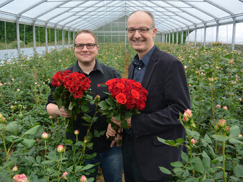 Zwei Männer mittleren Alters halten zwei Sträuße mit roten Rosen in den Händen. Sie tragen beide eine Brille und strahlen.