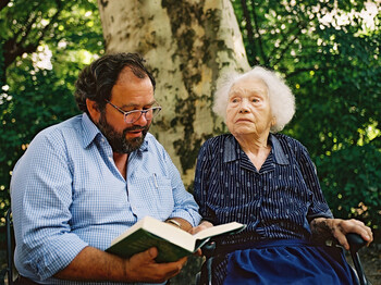 Ein Mann mittleren Alters sitzt neben einer alten Dame. Die Dame trägt eine blaue Kleidung und sitzt im Rollstuhl. Der Herr liest ihr etwas aus einem dicken Buch vor und die Dame hört ihm gespannt zu.