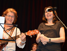 Eine Frau mittleren Alters spielt auf einer Gitarre. Eine Frau neben ihr singt und lacht.