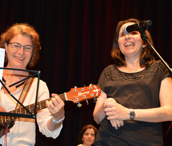 Eine Frau mittleren Alters spielt auf einer Gitarre. Eine Frau neben ihr singt und lacht.