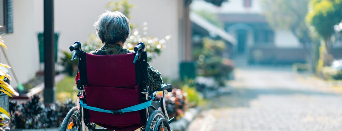 Workshop: Achtsame Begleitung von Menschen mit Demenz Eine im Rollstuhl sitzende Seniorin befindet sich in einer Hauseinfahrt und schaut die Straße hinunter.
