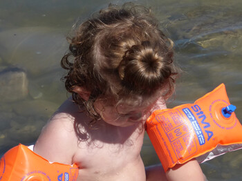Ein kleines Kind mit orangen Schwimmflügeln kühlt sich im seichten Wasser ab. Die braunen Haare hat es mit einem Dutt nach oben zusammengebunden.  
