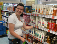 Simone Kneringer, eine junge, erwachsene Frau mit einem weißen Top ist in einem Drogerkiemarkt am Regale Einräumen. Sie lächelt dabei in die Kamera. 