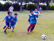 Eine Gruppe junger Buben, die beim Fußballspielen sind. Ein Junge will gerade den Ball schießen, die anderen laufen ihm hinterher.