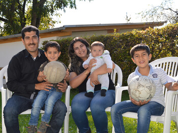 Eine junge Familie mit drei Kindern sitzt in einem Garten auf Plastikstühlen. Sie wirken glücklich und zwei der jungen Buben halten einen Fußball in der Hand. 