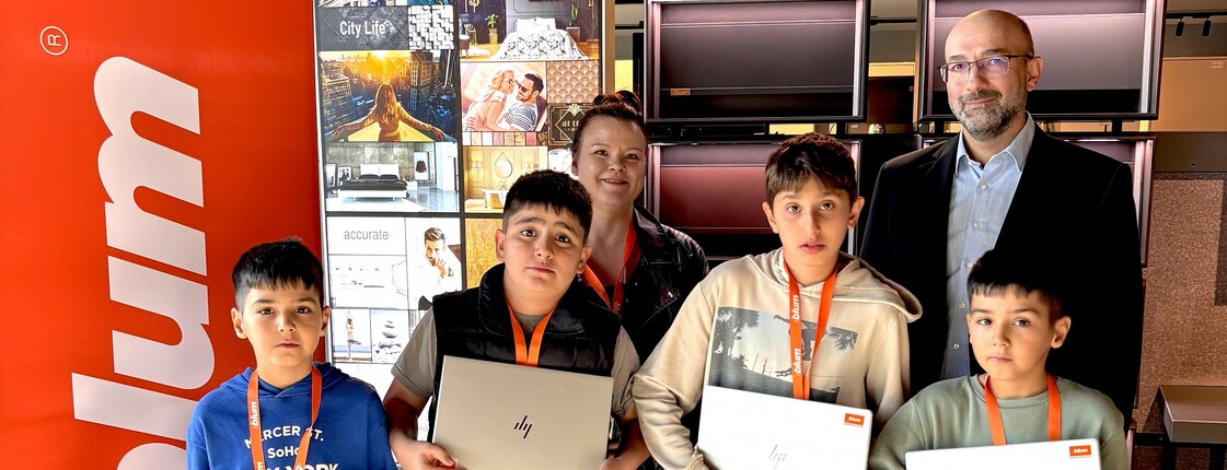 Blum-Mitarbeiter mit Kindern und Laptops vor Firmenwand