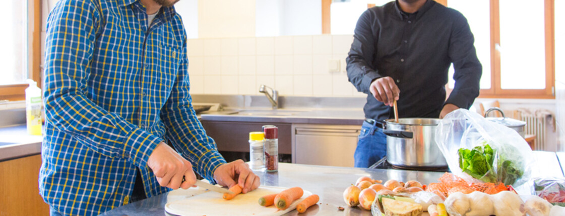 Zwei männliche Flüchtlinge mittleren Alters sind in einer großen und gut ausgeläuteten Küche am Kochen. Ein Mann schneidet gerade Karotten in kleine Stücke, der andere rührt mit einem Kochlöffel das Gericht in einem hohen Kochtopf um. Beide tragen ein blaues bzw. ein schwarzes Hemd. Ihre Haare sind schwarz. 