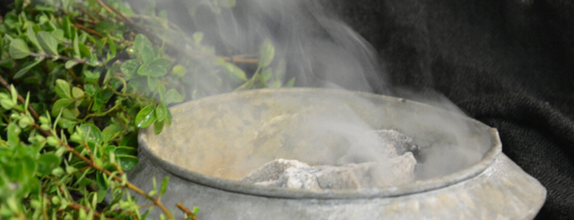 Das Bild zeigt einen grauen Kelch, aus dem Rauch kommt. Neben dem Kelch ist ein grüner Busch.