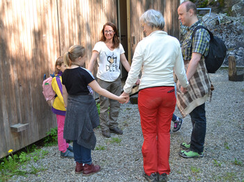 Drei Kinder stehen mit drei Erwachsenen in einem Kreis im Freien zusammen. Dabei halten sich alle am Arm.