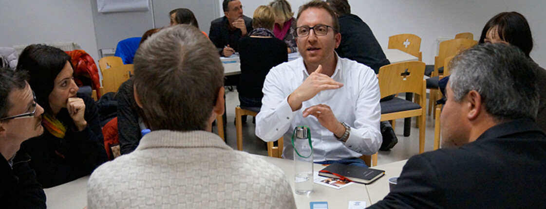 Im Vordergrund ist ein Tisch zu sehen, an dem Menschen im Gespräch sind. Ein Mann mit Brille und weißem Hemd gestikuliert, die anderen hören zu. Im Hintergrund sind weitere Tisch-Runden mit Menschen im Gespräch zu sehen.