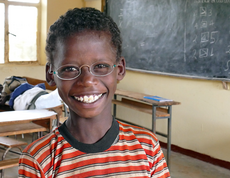 Ein kleiner Junge sitzt in einer Schulklasse. Er trägt eine Brille mit hellen Gläsern und strahlt. 