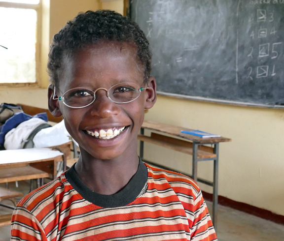 Ein kleiner Junge sitzt in einer Schulklasse. Er trägt eine Brille mit hellen Gläsern und strahlt. 