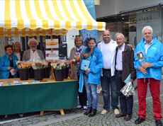 Ein Marktstand mit vielen Menschen versammelt sich für ein Foto. Sie verkaufen bunte Rosen für einen guten Zweck.