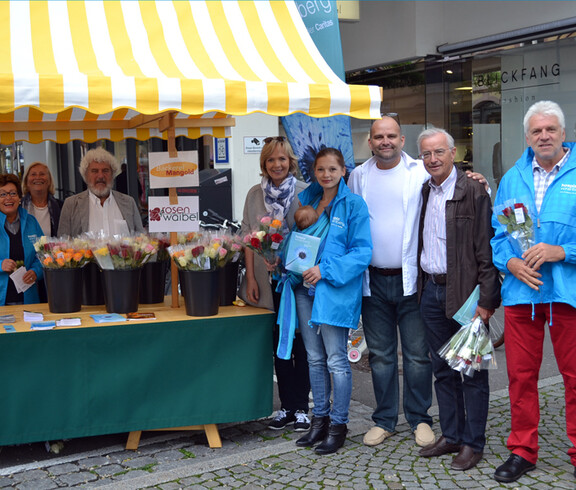Ein Marktstand mit vielen Menschen versammelt sich für ein Foto. Sie verkaufen bunte Rosen für einen guten Zweck.
