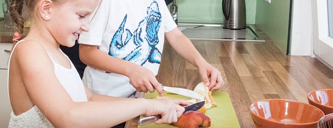 Zwei Kinder bereiten unter Anleitung einer Betreuungsperson Obst in einer Küche zu. Gemeinsames Kochen fördert Selbstständigkeit und Alltagskompetenzen.