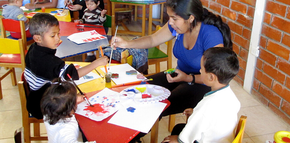 Viele Kinder, die gerade mit Wasserfarben malen. Die Kinder und Betreuerinnen sitzen auf bunten Stühlen und malen auf bunten Tischen.