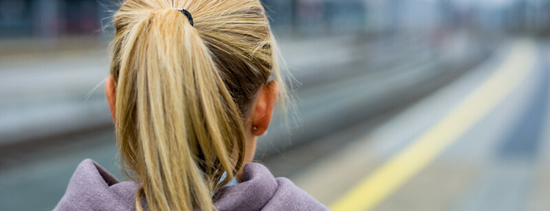 Eine Jugendliche mit langen blonden Haaren, die sie zusammengebunden hat, steht an einem  Bahnhof und blickt in die Ferne. Bis auf sie ist das Bild unscharf. Man sieht nur ihr Rücken. Sie trägt einen grauen Pullover. 