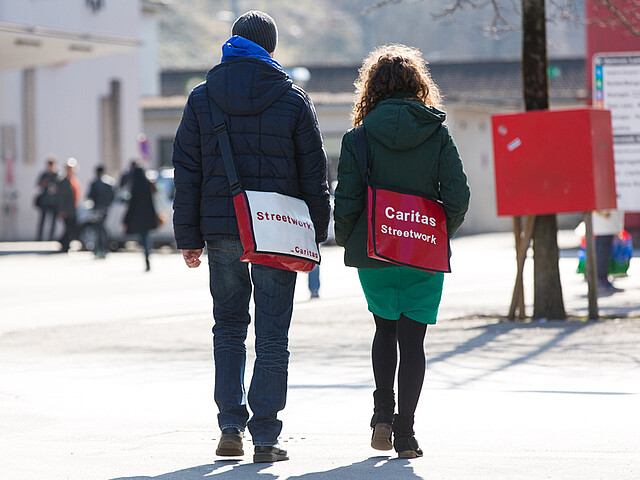 Zwei junge Erwachsene, eine Frau und ein Mann, spazieren Richtung Bahnhof Feldkirch. Sie wurden von hinten fotografiert. Beide tragen sie ein Umhängtasche auf denen jeweils "Streetwork" und "Caritas" steht. Beide sind sie in Winterkleidung angezogen. 