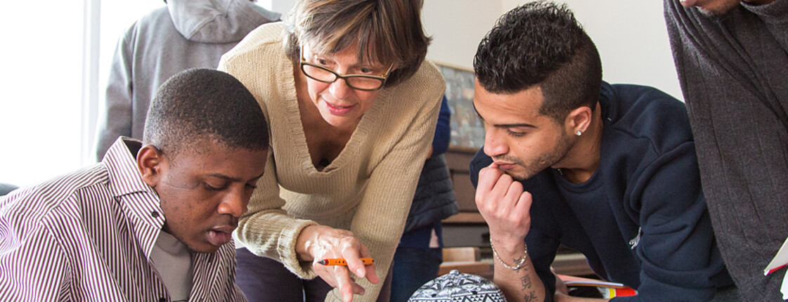 Flüchtlinge aus Afrika und dem nahen Osten lernen Deutsch. Sie sitzen gespannt und konzentriert an einem Tisch und blicken auf ihre Unterlagen. Eine Lehrerin hilft ihnen dabei den Stoff besser aufzunehmen und steht für jede Frage offen. 