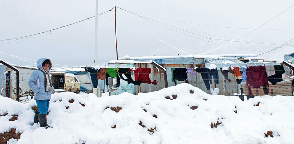 Ein kleines Kind steht auf einem Schneehaufen. Es ist sehr warm angezogen. Im Hintergrund sind Fahrzeuge, Wäscheleinen und Hütten.