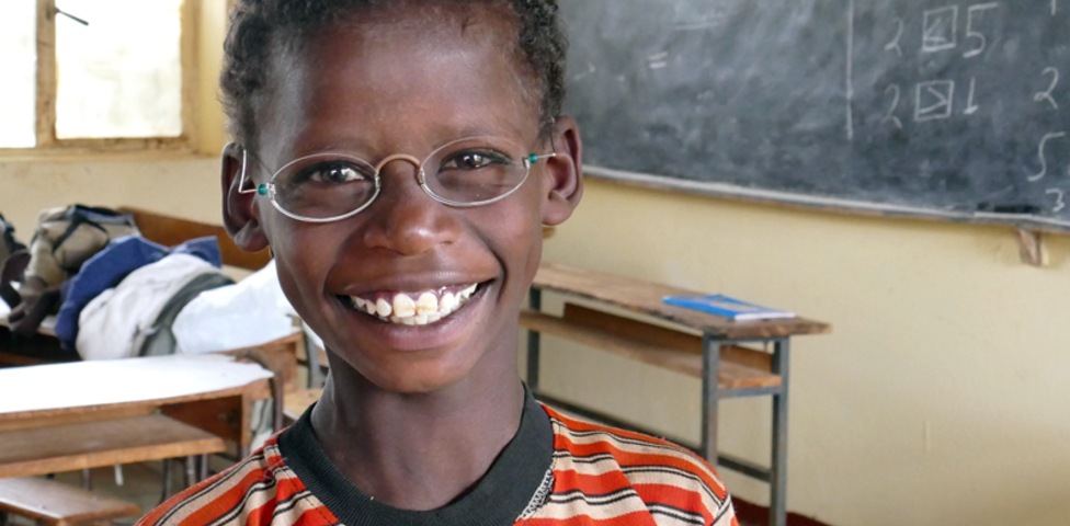 Ein kleiner Junge sitzt in einer Schulklasse. Er trägt eine Brille mit hellen Gläsern und strahlt. 