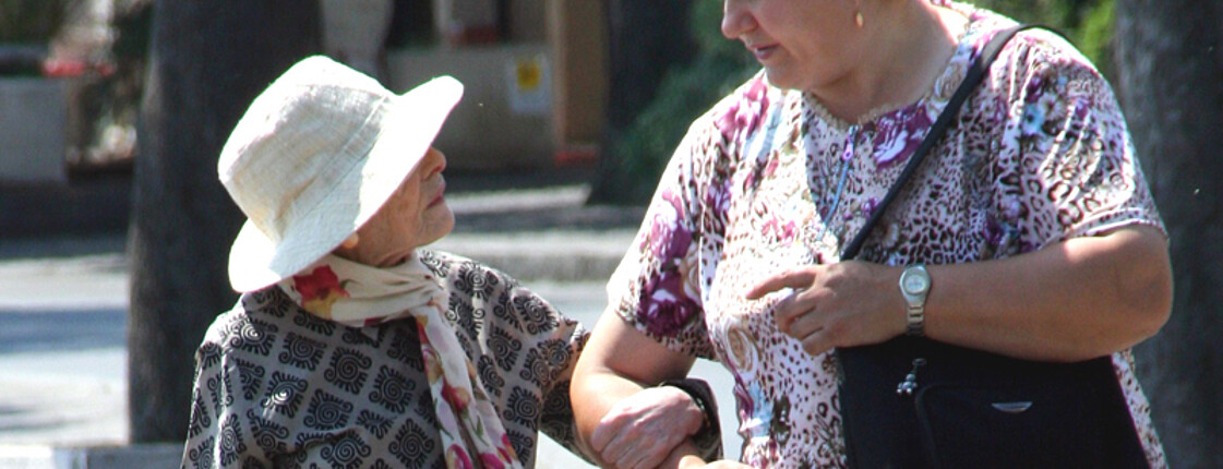 Eine ältere kleine Frau mit Sonnenhut wird von einer großen Frau mittleren Alters durch die Stadt geführt. Die jüngere Frau hat eine schwarze Handtasche. 