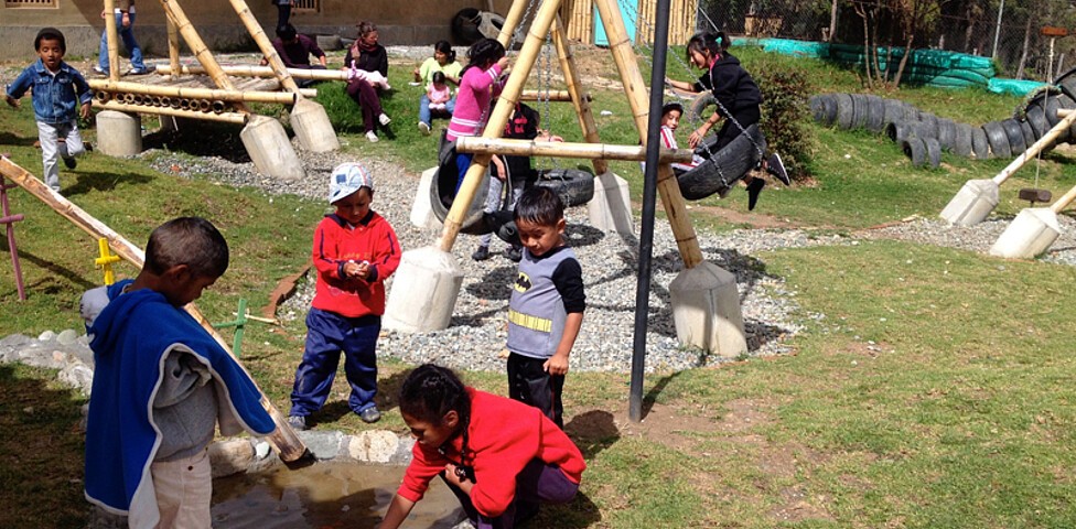 Ein Spielplatz im Freien, auf dem zahlreiche Kinder spielen. Manche sind beim Schaukeln, andere spielen mit dem Wasser. 
