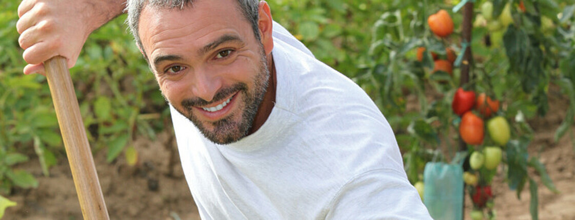 Ein Asylwerber mittleren Alters wird durch die Nachbarschaftshilfe beschäftigt. Er ist in einem Garten, hält einen Rechen in der Hand und lächelt in die Kamera. Er hat ein schlichtes weißes t-Shirt und eine Jeans an. Im Hintergrund sind Tomatenstöcke. 
