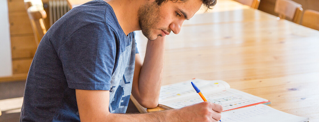 Ein junger erwachsener Flüchtling aus dem Nahen bzw. Mittleren Osten lernt an einem großen Hölzernen Tisch Deutsch. Dabei verwendet er ein Übungsbuch und einen Notizblock. Er hat kurze braune lockige Haare und trägt ein dunkelblaues t-Shirt. 