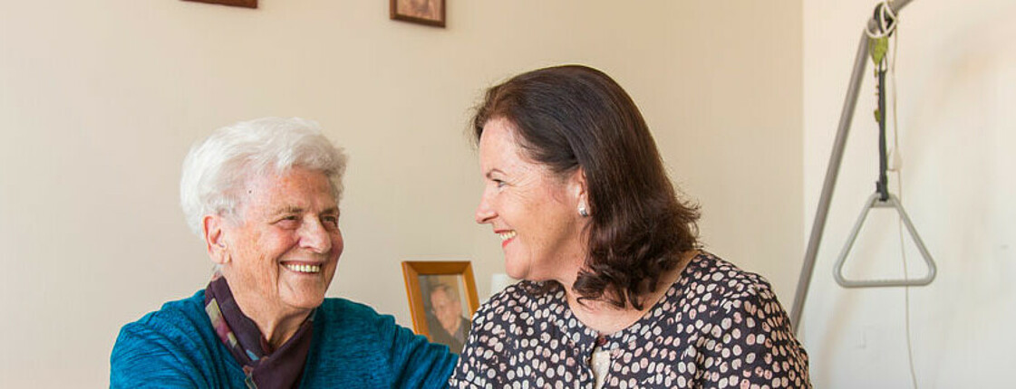 Zwei Frauen, die eine im mittleren, die andere im höheren Alter, schauen einander ins Gesicht. Die ältere der beiden Frauen hat graußes Haar, während die andere gefärbte Haare trägt. Beide sind erfreut und lachen. 