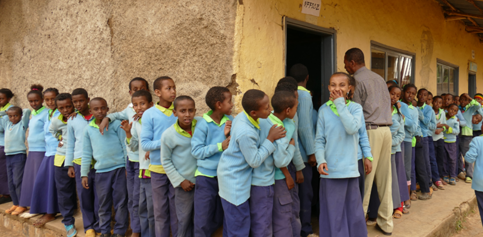 Viele Schulkinder stehen vor einer Schule. Alle tragen eine blaue Uniform. Die Kinder wirken sehr fröhlich. Ein Mädchen lacht und hält sich die Hand vor den Mund. 