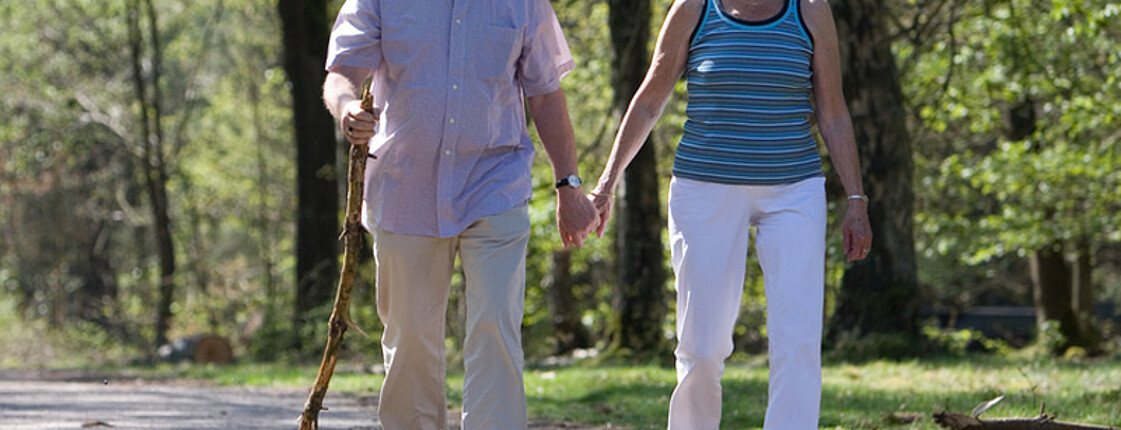 Zwei ältere Personen sind im Wald am Spazieren. Der Mann trägt ein weißes Hemd und eine beige Hose und die Frau eine weiße Hose und ein blaues Top. Der Mann hält zusätzlich einen Stock in der Hand. 