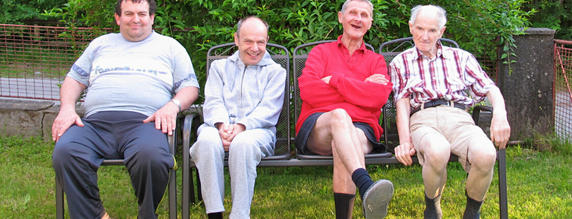 Vier Männer sitzen im Garten auf schwarzen Stühlen. Sie sind teils mittleren Alters teils schon Pensionisten. Sie fühlen sich in ihrer Umgebung wohl. 