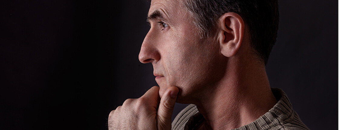 Ein Mann mittleren Alters wurde im Profil fotografiert. Er hat mattschwarze kurze Haare und trägt einen hellbrauenen dicken Pullover. Seine linke Hand hält er an sein Kinn. 