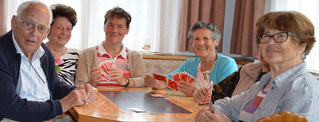 Seniorenerholung: Senioren spielen an einem Tisch karten miteinander. Sie sitzen alle dabei auf einer Sitzbank. 