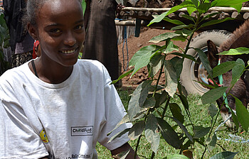 Ein junges Mädchen hält einen Mangobaum in den Händen und strahlt. Sie hat dunkles Haar und befindet sich auf einer Wiese.