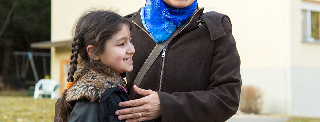 Zwei Flüchtlinge aus dem Nahen bzw. Mittleren Osten, eine Mutter mit ihrer kleinen Tochter, stehen vor einem Flüchtlingsheim. Sie sind beide schlicht und warm bekleidet. Die Mutter trägt neben einer dicken Jacke auch eine blaue Kopfbedeckung. Beide lachen dabei und sind glücklich. 