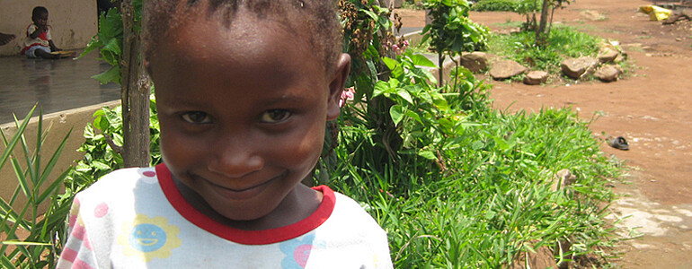Ein kleines dunkelhäutiges Mädchen in Mosambik trägt ein buntes T-shirt mit einem Blumenmuster und grinst verschmitzt in die Kamera.