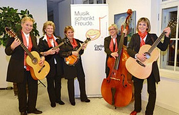 Fünf ältere Musikerinnen stehen mit ihren jeweiligen Instrumenten zu einem Gruppenfoto zusammen. Sie sind alle modisch und schick gekleidet. 